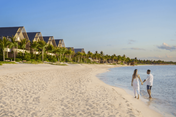 couple hold hands while walking on beach next to hotel villas